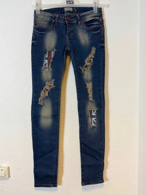 Blå slitna skinny jeans med flaggdetaljer - Midjan 35  cm  Längd 94 cm  Benvidd 22 cm 