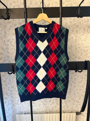 Rutig stickad väst från Cappuccini - Snygg stickad väst från Cappuccini med klassiskt rutigt argylemönster i marinblått, rött, grönt, vitt och gult. Västen har v-ringning och ribbade kanter, perfekt för att lyfta din outfit med en preppy touch.
