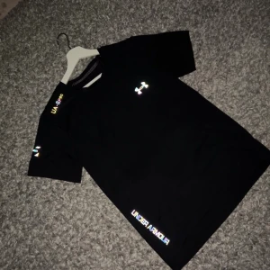 Svart Under Armour t-shirt - Snygg svart t-shirt från Under Armour med reflekterande logga på bröstet och textdetaljer på ärmen och nederkanten. Klassisk passform och korta ärmar, perfekt för träning eller chill. Tillverkad i mjukt och lätt material som andas.