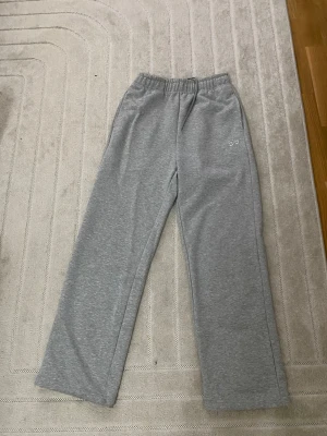 Grå mjukisbyxor från Alo - Snygga grå mjukisbyxor från Alo med broderad logga på ena benet. Byxorna har rak passform, resår i midjan och dragsko för extra komfort. Perfekta för en chill och avslappnad stil. Tillverkade i mjukt material som känns skönt mot huden.