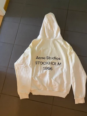 Vit hoodie Acne Studios Stockholm - Snygg vit hoodie från Acne Studios med tryck på ryggen där det står 'Acne Studios STOCKHOLM 1996'. Hoodien har huva och ribbade muddar vid ärmslut och nederkant. Perfekt för dig som gillar minimalistisk och clean stil.
