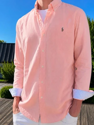 Ljusrosa skjorta från Ralph Lauren - Färgen är KORRAL FÄRGAD. (Moddelen är 185 och väger 76KG Klassisk ljusrosa skjorta från Ralph Lauren med button-down krage och broderad logga på bröstet. Skjortan har långa ärmar med randiga manschetter och vita knappar framtill. Perfekt för en stilren och avslappnad look.