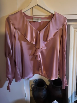Rosa volangblus från Neo Noir - Supersöt rosa blus från Neo Noir med volangdetaljer längs den djupa v-ringningen och framtill. Blusen har långa ärmar och är gjord i ett glansigt, mjukt tyg som ger en lyxig känsla. Perfekt för dig som vill ha en feminin och trendig look.