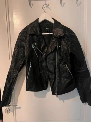 Svart biker skinnjacka med dragkedjor - Snygg svart skinnjacka i klassisk bikerstil med snedställd dragkedja och flera silverfärgade dragkedjor framtill. Jackan har bred krage och markerade axlar för en cool look. Perfekt för dig som vill ha en edgy stil.