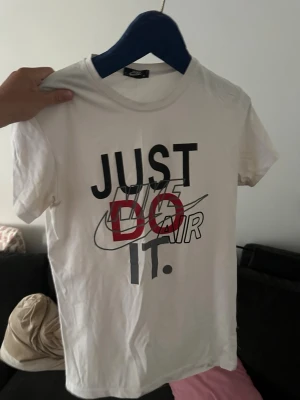 Vit Nike t-shirt med tryck - Säljer en vit t-shirt från Nike med det klassiska 'Just Do It' trycket i svart och rött framtill. T-shirten har korta ärmar och rund hals. Perfekt för en sportig och avslappnad stil.