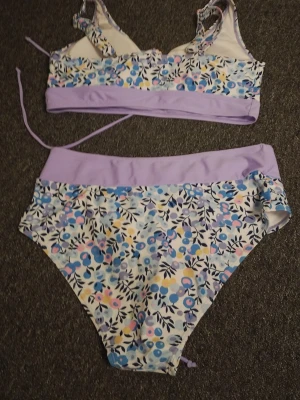 Ljuslila blommig bikini med hög midja - Söt bikini med hög midja i ljuslila och blommigt mönster i blått, gult och rosa. Toppen har smala axelband och knyt i ryggen för justerbar passform. Både överdel och underdel har breda ljuslila kanter. Perfekt för strandhäng i sommar!