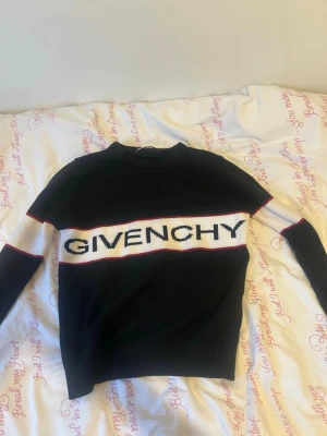 Svart stickad tröja från Givenchy - Snygg svart stickad tröja från Givenchy med vit panel och stor logga över bröstet. Tröjan har rund hals, långa ärmar och röda detaljer längs panelen. Använder inte längre då jag har bytt still.