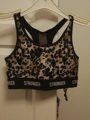 Leopardmönstrad sport-bh från Stronger - Sport-bh från Stronger med leopardmönster i beige, svart och brunt. Bred resår med loggan 'STRONGER' i vitt runt om. Racerback-modell med meshdetaljer på ryggen för extra ventilation. Perfekt för träning och gym. Kan tvättas innan jag skickar och kan mötas upp om det är i Karlstad.