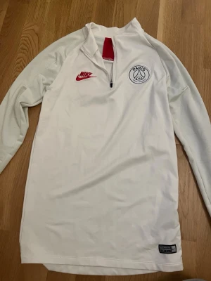 Vit Nike PSG träningströja - Snygg vit långärmad träningströja från Nike med PSG-logga på bröstet och röd Nike-swoosh. Tröjan har halv dragkedja 