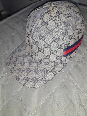 Gucci keps med GG-mönster - Snygg keps från Gucci i beige med klassiskt GG-monogram över hela kepsen. På sidan finns en röd och marinblå rand som ger extra lyxig känsla. Kepsen har böjd skärm och är tillverkad i canvasmaterial.