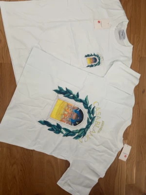 Vit Casablanca t-shirt med tryck - Vit t-shirt från Casablanca med färgglatt tryck framtill som visar en båge och gröna blad. T-shirten har rund hals och korta ärmar. Perfekt för dig som gillar stilrena plagg med unika detaljer.