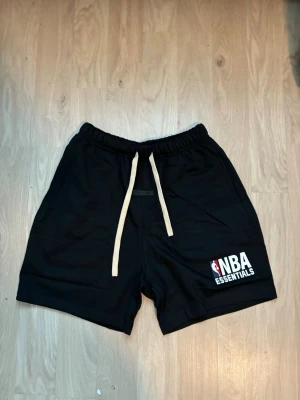 Svarta NBA Essentials shorts - Snygga svarta shorts från NBA Essentials med vit snörning i midjan och tryckt logga på benet. Perfekta för dig som gillar basket och vill ha en sportig look. Mjukt material och avslappnad passform.