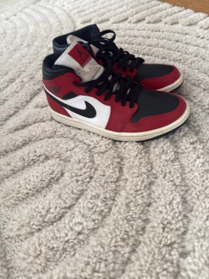 Air Jordan 1 Mid röd/svart/vit - Säljer ett par Air Jordan 1 Mid sneakers i klassisk röd, svart och vit färgkombination. Skorna har svart tåparti, röda paneler och vit sula samt sidopanel med svart swoosh. Tillverkade i läder med svarta skosnören och ikonisk Jumpman-logga på plösen. Pris kan diskuteras 