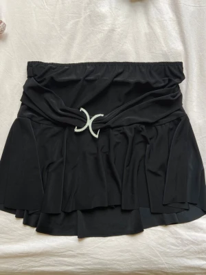 Svart kortkjol med glittrigt spänne - Supersnygg svart kortkjol med volang och ett glittrigt, dekorativt spänne framtill. Kjolen har en omlottliknande midja och är tillverkad i ett mjukt, stretchigt material som ger en skön passform. Perfekt för dig som gillar trendiga detaljer och vill sticka ut. Inbyggda shorts