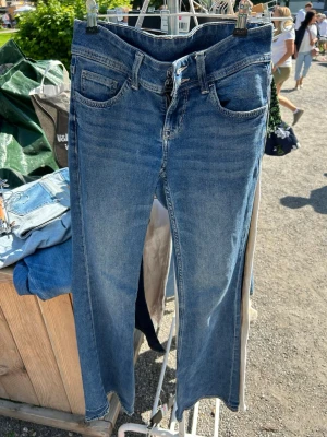 Blå bootcut jeans från Divided H&M - Säljer ett par klassiska blå jeans från Divided H&M i rak modell. Jeansen har fem fickor, låg midja midja och råa kanter vid bensluten för en trendig look. Tillverkade i denim med snygg tvätt och subtila slitningar. Perfekta till vardagsoutfits! Liten defekt som är lagad på baken som man ser på sista bilden