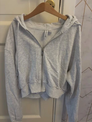 Ljusgrå hoodie med dragkedja - Snygg ljusgrå hoodie med dragkedja framtill och huva. Modellen har långa ärmar, ribbade muddar och en avslappnad passform. Perfekt för en chill och bekväm stil. Materialet är mjukt och skönt bomullsblandat tyg.