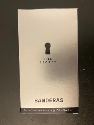 The Secret parfym av Banderas - The Secret från Banderas är en Eau de Toilette i en stilren silverfärgad förpackning med svart text och nyckelhålsdetalj. Flaskan rymmer 100 ml och är tillverkad av 20% återvunnet glas. Parfymen har en vegansk formula och består till 96% av ingredienser med naturligt ursprung. Den är helt ny och oanvänd.