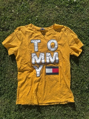 Gul Tommy Hilfiger t-shirt - Gul t-shirt från Tommy Hilfiger med stora vita bokstäver och logga på framsidan. Baksidan har också tryck med märkesnamnet i vitt. Klassisk rund hals och korta ärmar. Tillverkad i mjuk bomull, perfekt för en avslappnad stil.