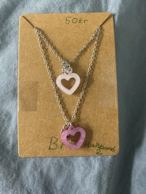 Sött halsband med hjärthängen - Tvådelat halsband med tunna silverfärgade kedjor och hjärtformade hängen i ljusrosa och lila plast. Perfekt för dig som gillar gulliga accessoarer och vill addera lite färg till din stil. Hängena har en öppen design och kedjorna är i metall.det e två bff halsband 