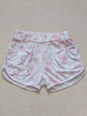 Rosa och vita mjuka shorts - Säljer ett par mjuka shorts i vitt med rosa batikmönster. Shortsen har elastisk midja, två fickor fram och rundade benslut. Perfekta för varma dagar eller att chilla i hemma. Barnstorlek, exakt storlek okänt.