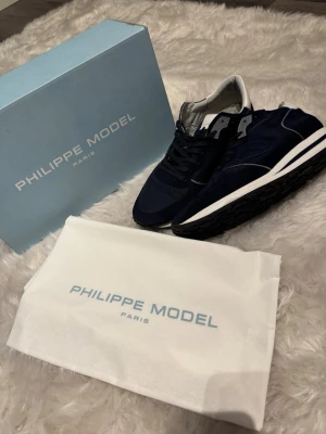 Marinblå Philippe Model sneakers - Snygga marinblå sneakers från Philippe Model med ovandel i mocka och mesh, vita detaljer och vit sula. Klassisk låg modell med svarta skosnören och diskret logga på sidan. Perfekta för en trendig och avslappnad stil.