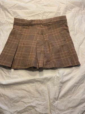 Brun rutig plisserad kjol från H&M - Säljer en brun rutig kjol med plisserad design och medel midja. Kjolen har bred linning och klassiskt rutmönster i beige och vitt. Perfekt för en trendig och preppy look.