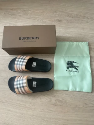 Burberry sandaler str 43 - Snygga sandaler från Burberry med klassiskt beige, svart och vitt rutmönster på remmen och svart sula. Tillverkade i syntetmaterial med märkets logga på sulan. Perfekta för en trendig och avslappnad stil.