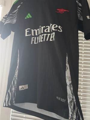 Arsenal svart matchtröja Adidas - Svart Arsenal matchtröja från Adidas med vita detaljer och grönt Adidas-märke. Tryck med 'Emirates Fly Better' på bröstet och klubbens röda logga. Sidorna har mönstrade paneler och materialet är lätt och andas, märkt HEAT.RDY.