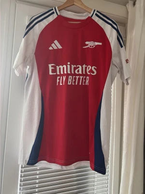 Arsenal matchtröja Saliba #2 - Snygg Arsenal matchtröja från Adidas i rött, vitt och blått med tryck 'Emirates Fly Better' på bröstet och 'Saliba 2' på ryggen. Tröjan har korta ärmar, klubbmärke på bröstet och ventilerande meshpaneler på sidorna. Perfekt för fotbollsfans!