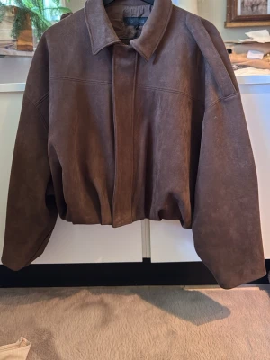 Brun oversized mockaimitations jacka  - Snygg brun mocka imitations jacka i oversized modell med breda axlar och klassisk krage. Jackan har dold dragkedja framtill och ett mjukt, matt skinn som ger en cool vintagekänsla. Perfekt för dig som gillar en avslappnad och trendig stil.