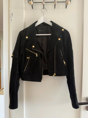 Svart mockajacka med guldiga detaljer - Snygg svart mockajacka med guldiga dragkedjor och knappar. Kort modell med flera fickor och coola detaljer på ärmarna. Perfekt för dig som vill ha en edgy look och gillar klassisk bikerstil. Det finns något märke samt smitning i tyget, se bild 
