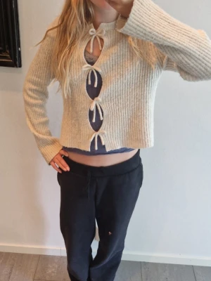 Beige stickad tröja med rosett, Kappahl - Beige stickad tröja från Kappahl med ribbad struktur och tre rosetter framtill. Kortare modell med långa ärmar och rund halsringning. Perfekt för lager på lager och en cool vardagslook.