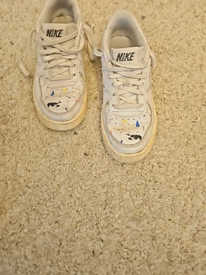 Nike vita sneakers med färgstänk - Unika vita Nike sneakers med färgstänk i blått, gult och svart. Klassisk låg modell med vit sula och grå swoosh på sidan. Skorna har snörning och är tillverkade i läder med detaljer i mocka. Perfekta för dig som vill sticka ut med en kreativ och personlig stil.
