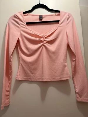Ljusrosa ribbad topp med rynk - Söt ljusrosa långärmad topp med ribbad struktur och rynkdetalj framtill. Toppen har en lätt v-ringad hals med diskret spetskant och är croppad i modellen. Perfekt för dig som gillar en feminin och trendig stil.