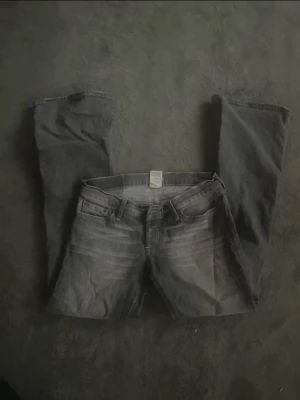 Grå low waist bootcut jeans - Säljer ett par jättesnygga low waist grå bootcut jeans, vit brodyr på bakfickorna. Jeansen har klassisk femficksmodell, bälteshällor och knappgylf. tyvvär har knappen gått av!🩷 midjemått- 39cm rakt över Innerbenslängd- 78cm Storlek-28 Pris kan diskuteras!