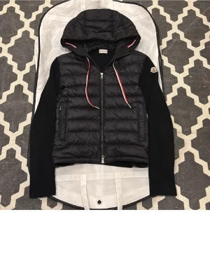 Svart Moncler cardiganjacka med huva - Svart cardiganjacka från Moncler med quiltad front, stickade ärmar och huva. Jackan har dragkedja, två fickor och röda/vita dragsnören. Ikonisk Moncler-logga på ärmen och snygg blandning av material för en sportig look. Ena drag kedjan e sönder på vänster sida kan även tänka mig byta 