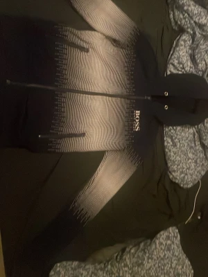 Svart hoodie från BOSS med mönster - Snygg svart hoodie från BOSS med dragkedja och vit grafisk gradient på ärmar och bröst. Märkeslogga på vänster sida av bröstet. Utrustad med huva och två fickor med dragkedja. Tillverkad i mjukt material, perfekt för en avslappnad streetwear-look.