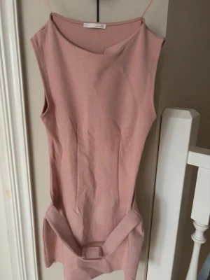Rosa ärmlös klänning från Zara - Säljer en stilren rosa klänning från Zara i ärmlös modell. Klänningen har en enkel, rak passform och ett matchande bälte i midjan som ger en snygg siluett. Perfekt för dig som gillar minimalistisk design och mjuka färger.