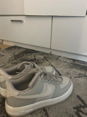 Nike Air Force 1 grå/vita sneakers - Snygga Nike Air Force 1 sneakers i grått och vitt. Klassisk låg modell med vit sula, perforerad tåbox och ikonisk Swoosh på sidan. Tillverkade i läder och textil med matchande grå skosnören. Perfekta för dig som gillar stilrena och tidlösa sneakers.