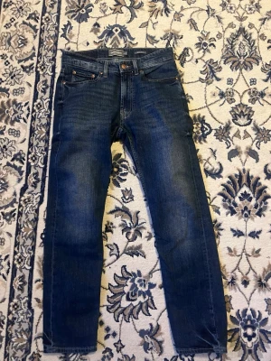 Mörkblå jeans från Dressmann - Säljer ett par mörkblå jeans från Dressmann med klassisk femficksmodell och raka ben. Jeansen har kopparfärgade nitar och knapp, samt diskreta slitningar för en snygg vardagslook. Materialet är mjukt denimtyg som sitter skönt hela dagen.