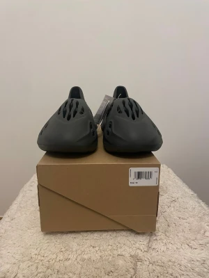 Svarta Yeezy Foam Runner carbon - Säljer ett par svarta Yeezy Foam Runner tofflor med futuristisk design och många ventilationshål. Tillverkade i ett lätt och flexibelt skummaterial som ger en unik look och skön känsla. Perfekta för dig som gillar streetwear och vill sticka ut.