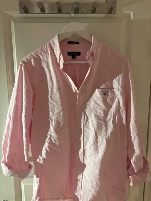 Ljusrosa oxfordskjorta från Gant - Klassisk ljusrosa oxfordskjorta från Gant med button-down krage och bröstficka med broderad logga. Skjortan har vita knappar och är tillverkad i bomull. Perfekt för en clean och stilren look.