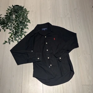 Svart skjorta från Ralph Lauren - Klassisk svart skjorta från Ralph Lauren med lång ärm och vit knappstängning. Skjortan har en röd broderad logga på bröstet och är tillverkad i bomull. Perfekt för en stilren och avslappnad look.