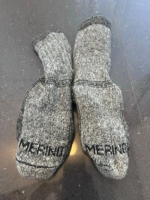 Grå melerade ullstrumpor från Merino - Ett par tjocka, grå melerade strumpor från Merino. De är stickade i ull och har svart text på tårna. Perfekta för kalla dagar och riktigt sköna att ha på sig. Klassisk modell med ribbad kant upptill och förstärkta partier vid häl och tå. Står inte någon storlek på, men uppskattar 36-38.