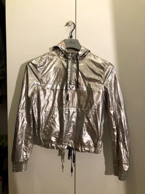 Metallic silver vindjacka med huva - Snygg metallic silver vindjacka med huva och dragkedja framtill. Jackan har svarta snören vid huva och midja, samt en stor magficka. Materialet är glansigt och ger en futuristisk vibe. Perfekt för dig som vill sticka ut i mängden.