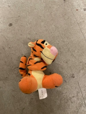 Tigergosedjur från Nalle Puh - Mjukisdjur som föreställer Tiger från Nalle Puh. Gosedjuret är orange med svarta ränder, ljusgul mage och rosa nos. Tillverkad i mjukt tyg och har en liten svans. Perfekt som samlarpryl eller present till någon som gillar Disney.