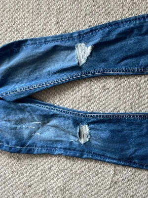 Blå ripped jeans med slitningar - HELT NYA ALDRIG ANVÄNDA! Ett par blå jeans i denim med tydliga slitningar och hål på benen för en trendig och avslappnad vibe. Jeansen har rak passform och klassisk blå tvätt. Perfekta för dig som gillar streetstyle och vill ha en edgy look.