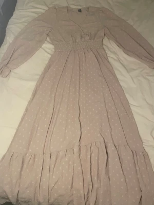 Ljusbeige långklänning med strukturmönster - Långklänning i ljusbeige ton med diskret strukturmönster. Klänningen har v-ringning, långa ärmar med resår vid handlederna och smock i midjan för en bekväm passform. Kjolen är volangig och tyget känns lätt och luftigt, perfekt för varmare dagar.