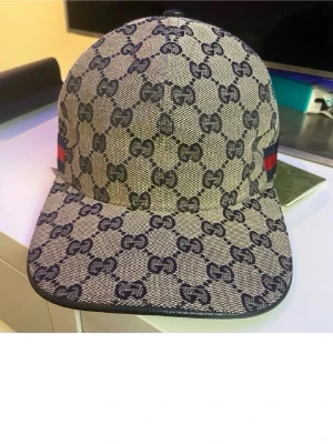 Gucci beige keps med GG-mönster - Säljer en beige keps från Gucci med klassiskt GG-monogram i mörkblått över hela kepsen. Kepsen har rak skärm och detaljer med röd och grön rand på sidan. Materialet är canvas med läderkant runt skärmen. Perfekt för dig som gillar exklusiva accessoarer. Helt ny oanvänd. Priset kan diskuteras 