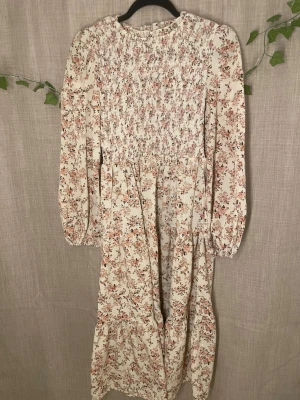 Blommig långklänning med smock - Romantisk långklänning i ljusbeige med småblommigt mönster i rosa och grönt. Klänningen har smockad överdel, långa ärmar med resår vid ärmslut och volangdetaljer i kjolen. Perfekt för vår och sommar.